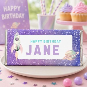 May include: A rectangular candy bar wrapper featuring a purple galaxy background with white speckles. The center white label displays the text HAPPY BIRTHDAY JANE in teal and purple. A rocket ship and a planet with rings are printed on the label.