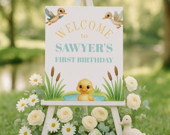 May include: A white rectangular sign on a wooden easel featuring the text WELCOME to SAWYER'S FIRST BIRTHDAY in yellow and light blue. The sign displays illustrations of three yellow ducklings, green reeds, and a small blue pond.
