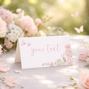 Pode incluir: Um cartão branco dobrado com o texto "your text" em escrita rosa, adornado com borboletas em aquarela e arranjos florais em tons de rosa e branco. O cartão está sobre um fundo suave e desfocado de flores.
