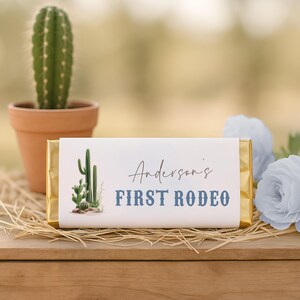 Può includere: Una barretta di cioccolato rettangolare avvolta in carta bianca con estremità in foglia d'oro. L'etichetta presenta un'illustrazione ad acquerello di un cactus e il testo "Anderson's FIRST RODEO" in corsivo nero e stampatello blu.