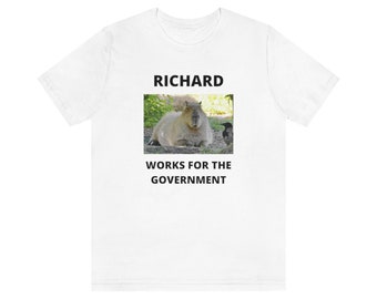 Puede incluir: Camiseta blanca con texto negro que dice "RICHARD WORKS FOR THE GOVERNMENT". La camiseta presenta una foto de un capibara y un cuervo.