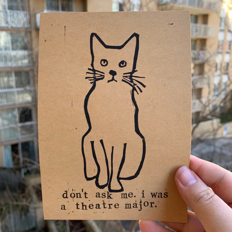 Pu&ograve; includere: Un'illustrazione in bianco e nero di un gatto seduto su uno sfondo marrone. Il testo "don't ask me. i was a theatre major." &egrave; stampato sotto il gatto.