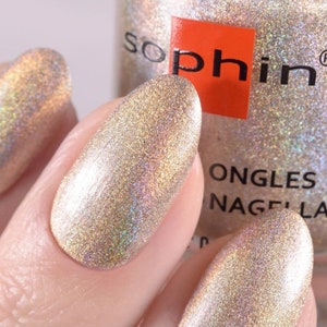 May include: A bottle of Sophi nail polish in a shimmery gold color with a holographic finish. The bottle is labeled "Sophi" and "Ongles Nagellack".
