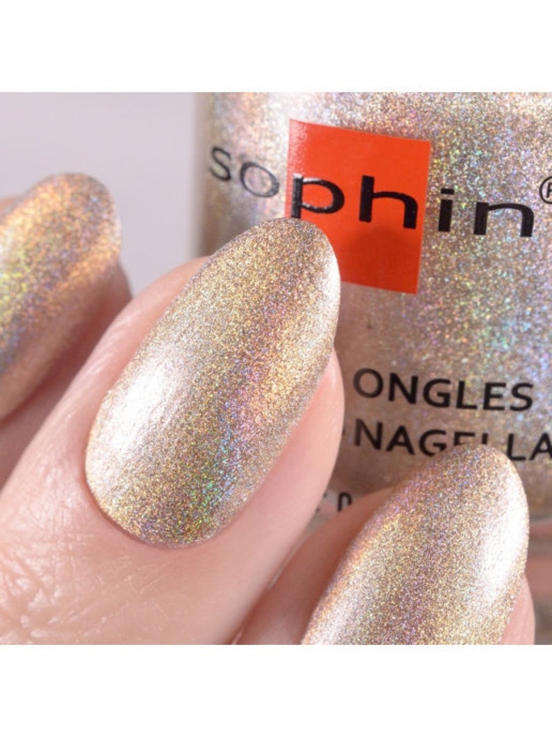 May include: A bottle of Sophi nail polish in a shimmery gold color with a holographic finish. The bottle is labeled "Sophi" and "Ongles Nagellack".