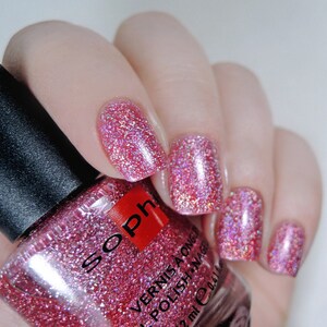 K&ouml;nnte beinhalten: Eine Flasche rosa Glitzer-Nagellack mit dem Markennamen "Soph" auf dem Etikett. Die Flasche wird teilweise von einer Hand mit rosa Glitzer-Nagellack auf den Fingern&auml;geln verdeckt.