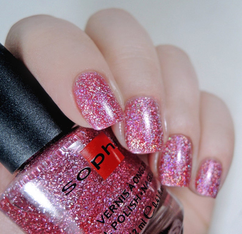 K&ouml;nnte beinhalten: Eine Flasche rosa Glitzer-Nagellack mit dem Markennamen "Soph" auf dem Etikett. Die Flasche wird teilweise von einer Hand mit rosa Glitzer-Nagellack auf den Fingern&auml;geln verdeckt.