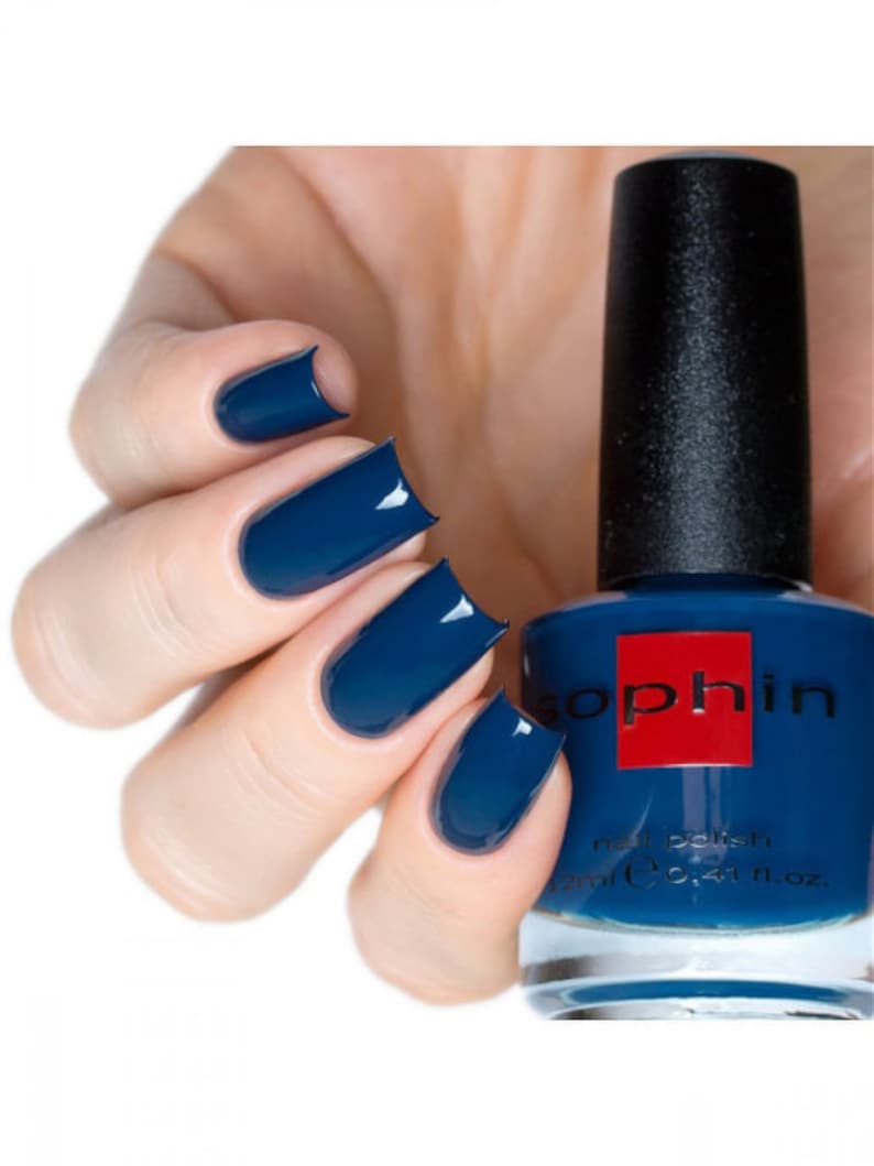 May include: A bottle of Sophiin nail polish in a deep blue color. The bottle is clear and shows the polish inside. A hand is shown with four fingernails painted with the same blue polish.