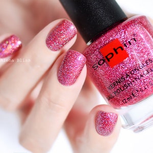 Puede incluir: Botella de esmalte de uñas rosa con una etiqueta cuadrada roja que dice "Sophin" y el texto "Vernis a Ongles Nail Polish Nagellack 12 ml e 0.4 fl oz". La botella está llena de esmalte de uñas rosa con purpurina. Se muestra una mano con esmalte de uñas rosa con purpurina en las uñas.