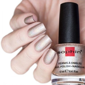 May include: A bottle of Sophiin nail polish in a shimmery champagne gold color. The bottle is 12 ml or 0.41 fl oz. The nail polish is shown on a hand with a light skin tone.
