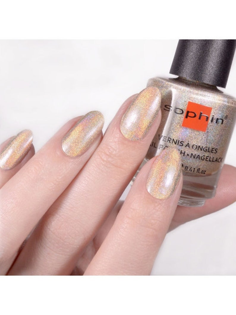 May include: A bottle of Sophiin nail polish in a shimmery gold color with a holographic finish. The bottle is labeled "Vernis &agrave; Ongles Nail Polish -  H.Nagellack 0.41 fl.oz."