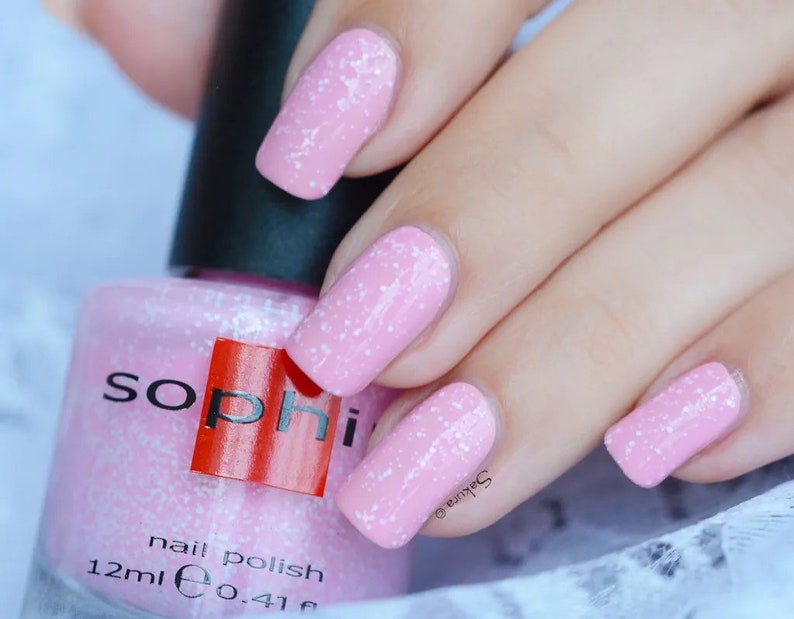 May include: Pink nail polish with a sparkly finish in a clear glass bottle with a black cap. The bottle label reads "soph" in red letters and "nail polish 12ml e 0.41 fl oz".