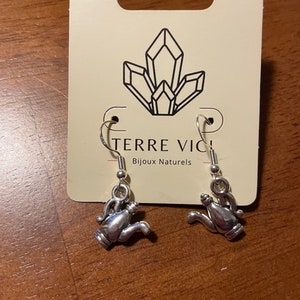 May include: Silver-colored teapot earrings with a hook closure. The earrings are displayed on a cream-colored card with the text "TERRE VICI Bijoux Naturels" and a crystal graphic. The earrings are on a wooden surface.