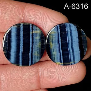 May include: Two round, polished gemstones with a striped pattern in shades of blue and yellow. The gemstones are held between fingers against a black background. The text "A-6316" is in the upper right corner.