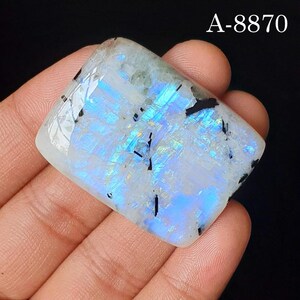 May include: A rectangular moonstone cabochon featuring a translucent white base with vibrant blue adularescence and scattered black tourmaline inclusions. The stone displays the text A-8870 in the upper right corner against a dark background.