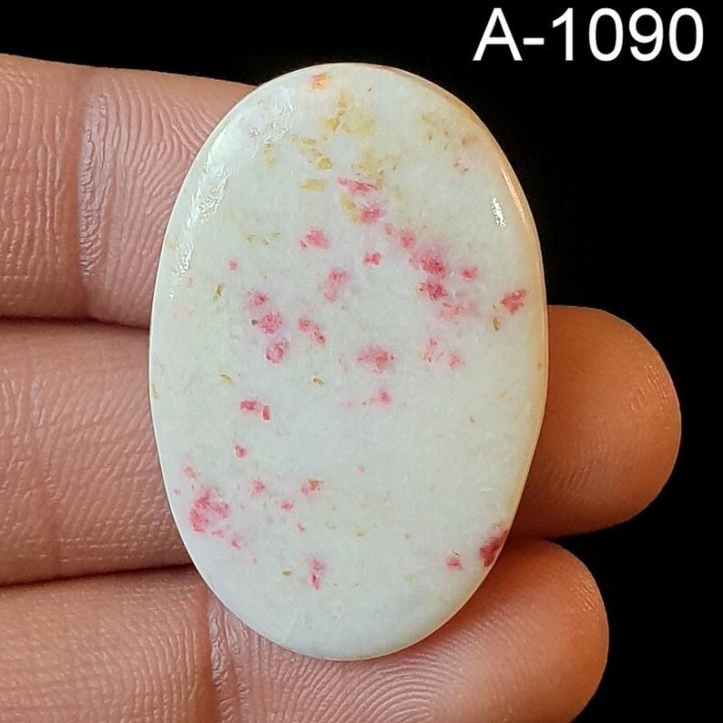 May include: A white oval cabochon with a speckled pattern of pink and yellow. The stone is approximately 2 inches long and 1.5 inches wide. A-1090