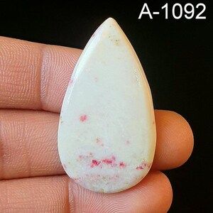 May include: A white teardrop-shaped cabochon with pink and red inclusions. The cabochon is approximately 2 inches long and 1 inch wide. A-1092