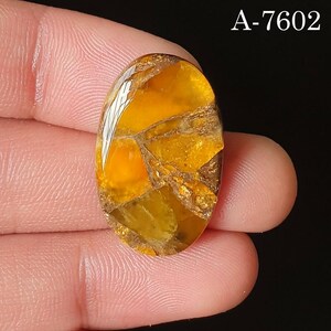 Puede incluir: Una piedra preciosa ovalada con un patrón de mosaico en tonos amarillo dorado, marrón y blanco. La piedra se sostiene entre los dedos, con el texto "A-7602" en la esquina superior derecha. La piedra preciosa tiene una superficie pulida y reflectante.