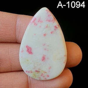 May include: A white cabochon gemstone with pink and yellow inclusions. The gemstone is in a teardrop shape and has a smooth, polished surface. A-1094
