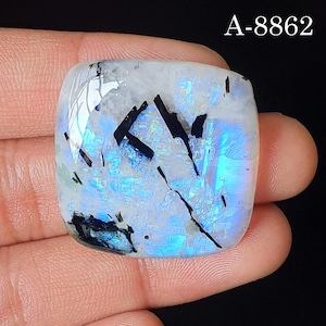 May include: A square-shaped polished moonstone cabochon featuring a translucent white base with vibrant blue adularescence and distinct black needle-like inclusions throughout the surface. The text A-8862 is printed in the upper right corner.