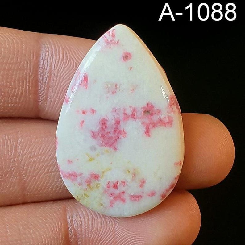 May include: A white cabochon with pink and yellow flecks. The cabochon is shaped like a teardrop and is approximately 2 inches long. The cabochon is labeled A-1088.