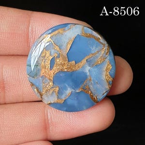 May include: A round, polished cabochon featuring a light blue base with intricate, branching metallic gold-colored veins throughout the surface. The text A-8506 is printed in white in the upper right corner of the image, identifying the item.