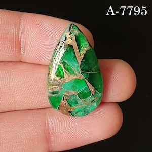 Könnte beinhalten: Ein tropfenförmiger Edelstein mit leuchtend grüner Farbe, durchzogen von goldfarbenen Adern. Der Stein ist poliert und reflektiert das Licht, wodurch ein mosaikartiges Muster entsteht. Der Text "A-7795" ist oben rechts sichtbar.
