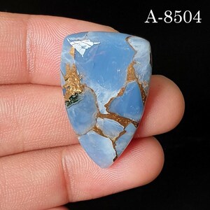 May include: A shield-shaped blue opal gemstone with a smooth, polished surface featuring intricate brown matrix veining throughout. The stone displays a light blue color with reflective highlights and is marked with the text A-8504 in the corner.
