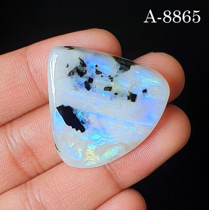 May include: A polished, triangular moonstone cabochon featuring a translucent white base with iridescent blue flashes and distinct black mineral inclusions. The stone displays the text A-8865 in the upper right corner against a dark background.