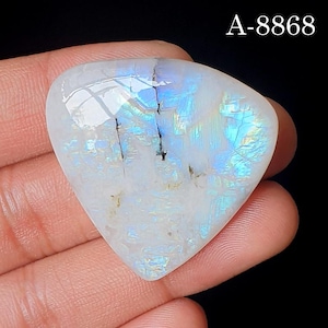 May include: A triangular moonstone cabochon with a smooth, polished surface displaying iridescent blue flashes and internal fractures. The stone features a white base color with visible light reflections and the text A-8868 in the top corner.