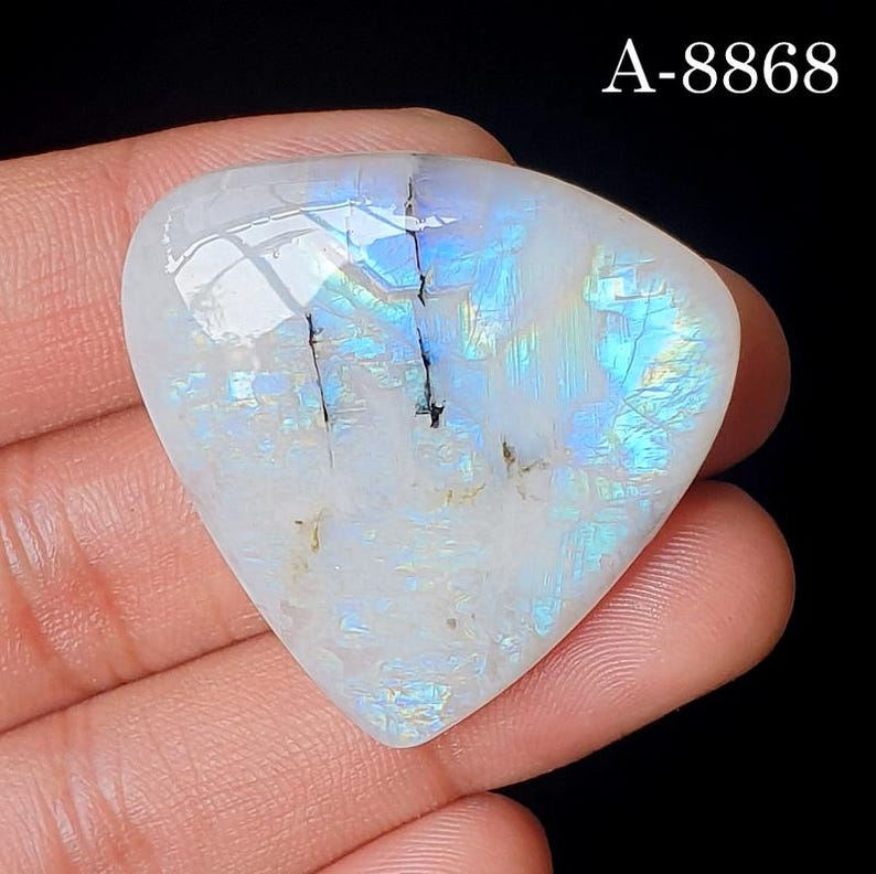 May include: A triangular moonstone cabochon with a smooth, polished surface displaying iridescent blue flashes and internal fractures. The stone features a white base color with visible light reflections and the text A-8868 in the top corner.