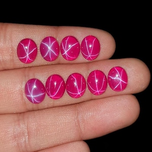 Puede incluir: Ocho gemas rosas ovaladas y vibrantes con efecto de estrella. Cada gema presenta un patrón de estrella blanca de seis rayos. Las gemas se muestran sobre un fondo oscuro.