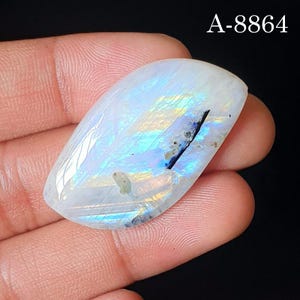 May include: A polished, oval-shaped moonstone cabochon featuring a translucent white base with iridescent blue and yellow flashes. The gemstone displays a distinct dark linear inclusion and small mineral specks. The text A-8864 is visible.