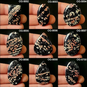 May include: Nine black and white gemstone cabochons with red and white inclusions. The cabochons are in a teardrop shape and are arranged in three rows of three. The cabochons are labeled with the numbers OG-8692 through OG-8700.