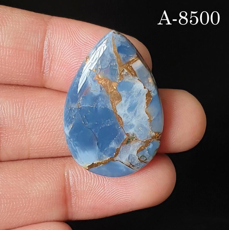 May include: A pear-shaped blue gemstone with a smooth, polished surface featuring intricate copper-colored veining throughout. The stone displays a mix of light and dark blue tones and is marked with the text A-8500 in the upper right corner.