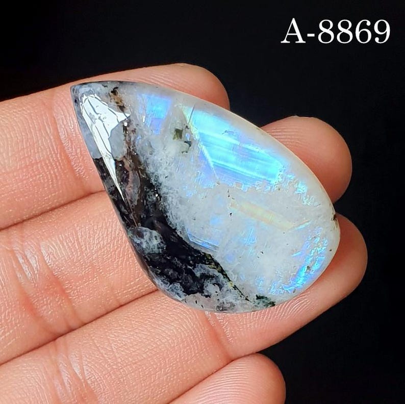 May include: A pear-shaped moonstone cabochon featuring a translucent white body with a distinct blue adularescence and dark black mineral inclusions. The stone displays a smooth, polished surface and is marked with the text A-8869 in the corner.