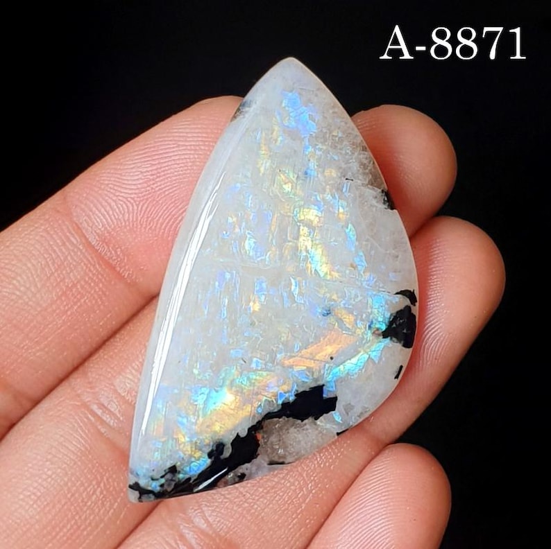 May include: A triangular moonstone cabochon featuring a polished surface with iridescent blue and yellow flashes, internal crystalline textures, and distinct black mineral inclusions. The text A-8871 is visible in the upper right corner.