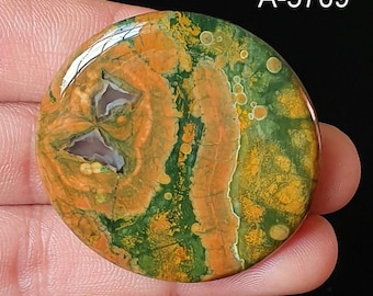May include: A round, polished gemstone with a unique pattern of orange, green, and white swirls. The stone has a natural, organic appearance, with the text "A-5709" in the upper right corner. The gemstone is held in a hand.