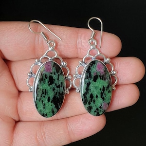 May include: A pair of oval-shaped dangle earrings featuring polished green and black zoisite stones with embedded magenta ruby inclusions. The stones are set in silver-toned metal frames with intricate filigree detailing and hook-style wires.