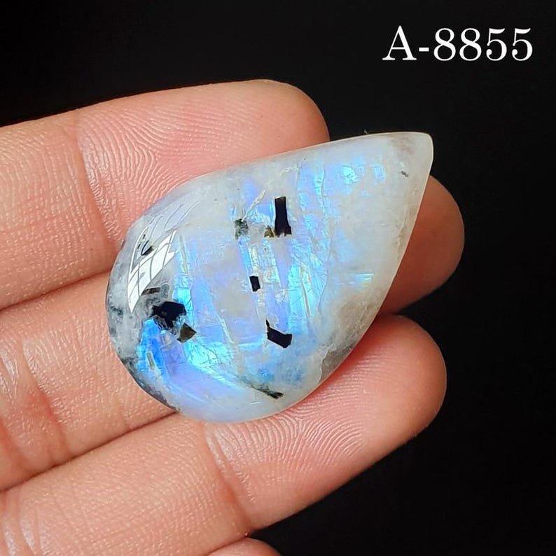 May include: A pear-shaped moonstone cabochon featuring a translucent white base with iridescent blue adularescence and several small, dark, rectangular inclusions. The stone is marked with the text A-8855 in the upper right corner.