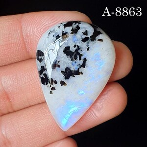 May include: A pear-shaped moonstone cabochon featuring a translucent white base with scattered black inclusions and a distinct blue adularescence shimmer. The stone is marked with the text A-8863 in the upper right corner of the image.