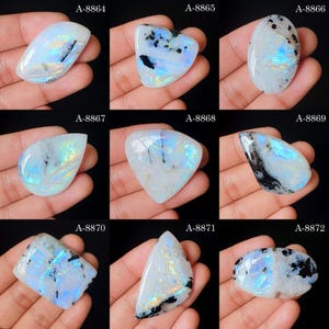 May include: A collection of nine polished moonstone cabochons with iridescent blue flashes and black inclusions, labeled A-8864 through A-8872. Each gemstone displays a unique shape, including oval, pear, and triangular forms with smooth surfaces.