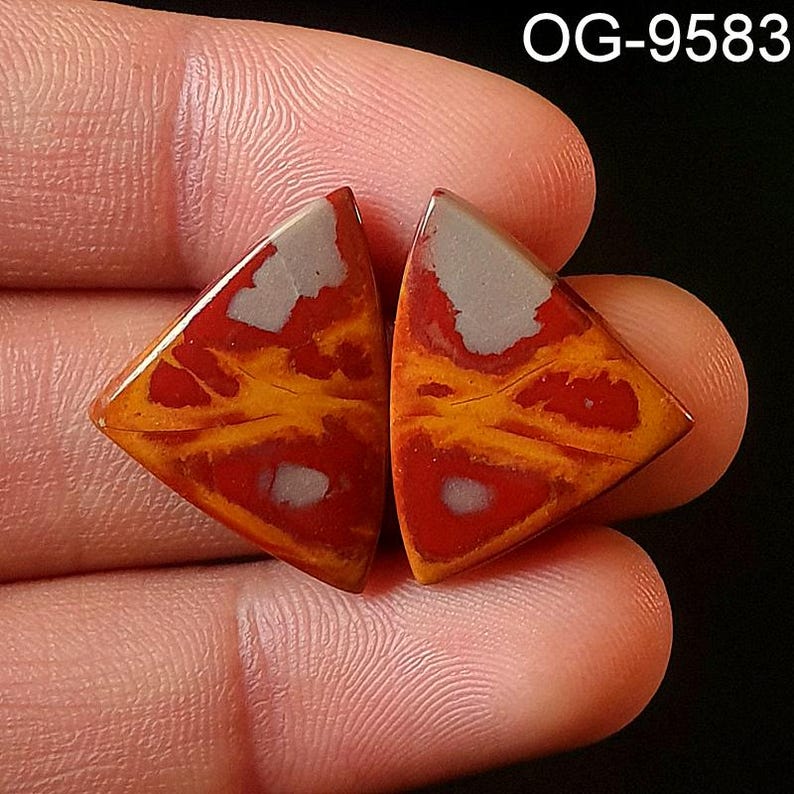 May include: Two triangular shaped red, orange, and white agate cabochons. The cabochons have a swirling pattern and are held in a hand.