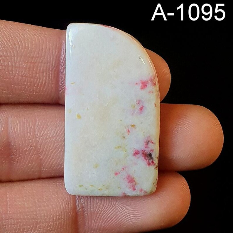 May include: A white cabochon with pink and yellow inclusions. The cabochon is shaped like a rectangle with rounded corners. The cabochon is approximately 2 inches long and 1 inch wide. A-1095