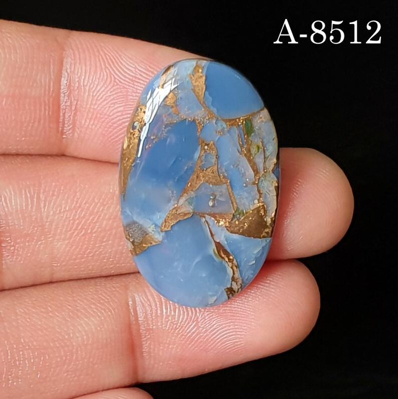 May include: An oval-shaped cabochon gemstone featuring a smooth, polished surface with a light blue base color and intricate, metallic copper-colored veining throughout. The text A-8512 is visible in the upper right corner of the image.