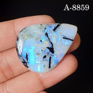 May include: A pear-shaped moonstone cabochon featuring a translucent white base with iridescent blue flashes and distinct black tourmaline needle inclusions. The stone is marked with the text A-8859 in the upper right corner against a black background.