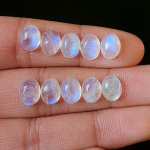 Puede incluir: Ocho piedras preciosas de forma ovalada, piedras lunares. Las piedras tienen un aspecto translúcido y lechoso con un sutil juego de luz azul y blanca. Las piedras se muestran sobre un fondo oscuro.