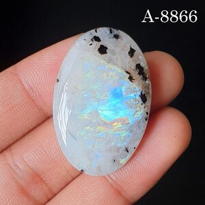 May include: An oval-shaped moonstone cabochon featuring a translucent white base with a vibrant blue and iridescent flash. The stone displays dark, irregular mineral inclusions near the top edge and is marked with the text A-8866 in the corner.