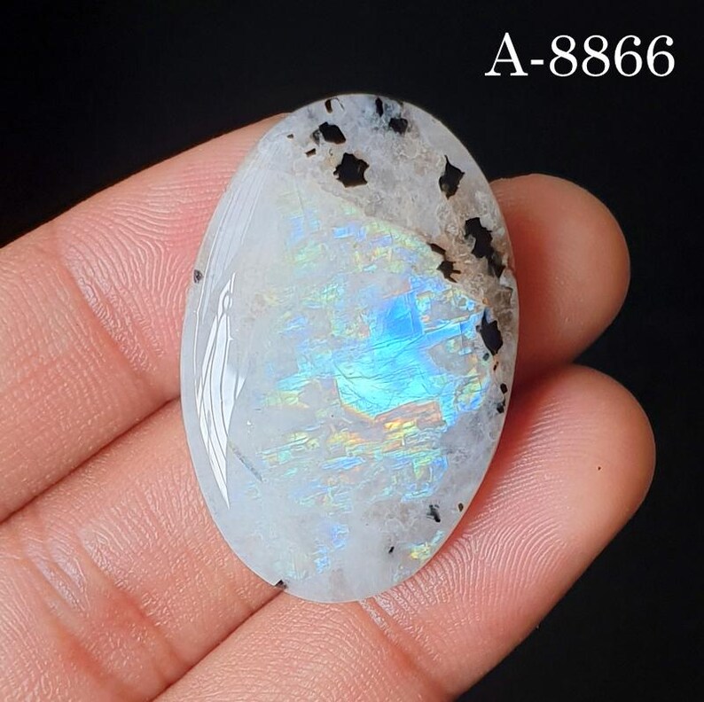 May include: An oval-shaped moonstone cabochon featuring a translucent white base with a vibrant blue and iridescent flash. The stone displays dark, irregular mineral inclusions near the top edge and is marked with the text A-8866 in the corner.