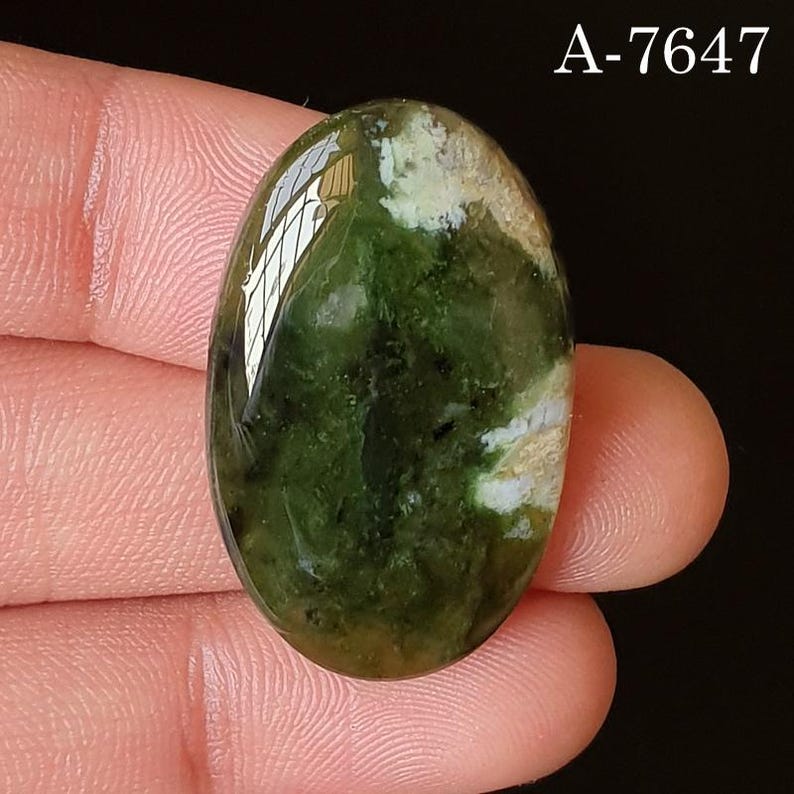 May include: Oval-shaped gemstone with a polished surface. The stone features a mix of green and white hues, with darker green areas and lighter, almost beige, patches. The text "A-7647" is visible in the upper right corner. The gemstone is held between fingers.