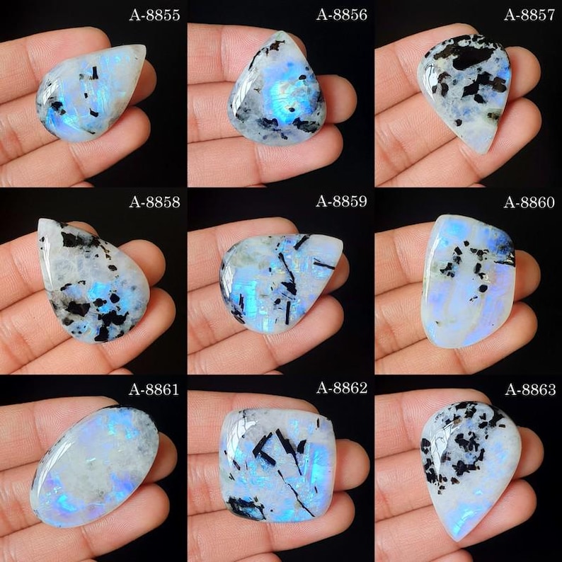 May include: A collection of nine polished moonstone cabochons, each featuring a unique iridescent blue adularescence and black inclusions. The stones are labeled A-8855 through A-8863, displaying various shapes including teardrop, oval, and square.